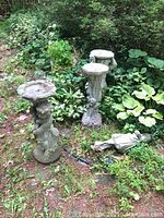 Three cement garden objects placed outdoors among grass and plants: two pedestals/statues and one planter. One statue is toppled, one shows a cherub figure, the planter rests on a stepping stone.