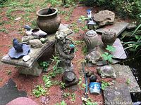 Wide view of outdoor garden setup with stone benches, large planter, child statue, resin fish fountain top, and various animal figures placed on and around stone benches and ground covered with pine needles.