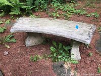Curved cement bench with two supporting legs, featuring decorative edges and natural weathering including dirt and hairline cracks, photographed outdoors in a garden setting.