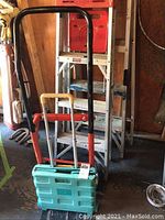 Photo showing black metal hand truck with wooden handle grips, teal rolling collapsible bin folded compact, and two metal frame step ladders stacked behind.