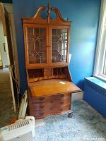 Full front view of vintage wooden secretary desk in a room with blue walls. Shows serpentine lower drawers and glass paneled doors on upper cabinet, and drop-down writing surface.