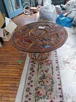 Top view showing the round carved wooden tabletop with detailed floral and leaf motifs carvings, positioned on a floral patterned rug indoors.