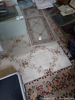 Photo showing large cream floral patterned rug partially visible with smaller rugs stacked on top and around it, with signs of wear and staining.