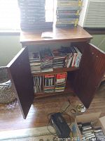 Open wooden cabinet filled with stacked CDs and DVDs, with an Optimus LSCT86 cassette deck and cables on the floor in front.