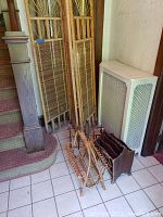 Photo shows three wood and rattan divider screen panels leaning against a staircase, next to a white heater. On the floor are two magazine racks: one rattan and one wooden with multiple compartments.