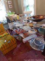 Wide view of wire shelving holding various glassware including bowls, dishes, and decorative items beside a striped yellow chair and window with lace curtains