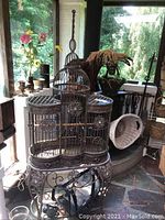 View from front showing the ornate metal bird cage on a decorative metal stand with floral embellishments, window background