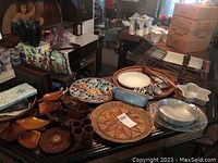 Photo of wood cups, napkin holders, carved trays, ceramic plates, metal racks, and warming tray arranged on a table.