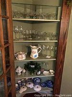 Wide side view of cabinet shelves showing a variety of glassware and china, including stemware, green glass, a pitcher, and cups.