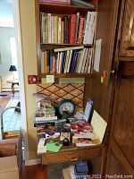 Wooden built-in shelving unit with multiple cookbooks on top shelves, drawers below holding various office supplies, pens, vintage ink well, tape rolls, and markers. Mechanical scale and untested clock present. Area shows visible wear and tear, packed densely.