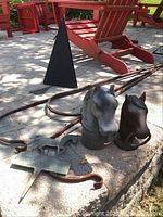 Photo of cast iron horse head post toppers in gray and black with metal rings, four long wrought iron shepherds hooks in rusty condition, and parts of a wind chime on concrete surface with red Adirondack chairs in background.
