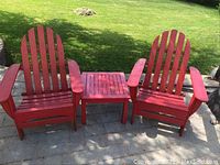 Front view of two red Adirondack chairs flanking a matching red side table on paved patio.