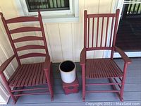 Two red painted wooden rockers, small red stool, and white ceramic crock on wooden porch floor
