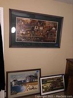 Three framed prints displayed on a wall: a large horizontal autumn village scene above two smaller prints: a coastal village beach scene and a Tudor-style cottage garden scene