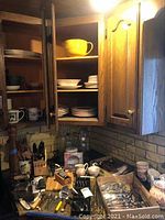 Kitchen corner with wooden cabinets open showing stacked plates, bowls, and other ceramic dishes; countertop holds metal flatware, utensils, and serving pieces in boxes and containers