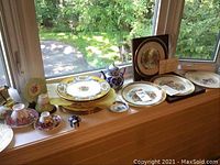 Wide shot of various plates and tea cups on a window sill including Lenox fox plates, Myotts plates and blue-and-white porcelain pieces.