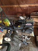 Full side view of rototiller showing engine, handle, wheels and blades lying on the ground of a cabin or shed.