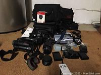 Full overview shot showing Nikon video camera recorder, Nikon camera with lens, lenses, Canon EVF-DC1 lens box, battery chargers, straps, and multiple camera bags in black fabric.