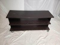 Front angled view showing dark brown wooden IKEA TV stand with two shelving tiers and caster wheels. Slight bend noticeable on the top shelf.
