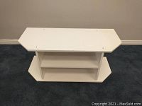 Front view of white TV stand or storage unit showing top, two shelves, and chamfered corners on rectangular base and top.