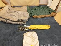 Beige tent fabric and green ground sheet laid flat on carpet. Metal poles bundled in yellow bag and beige carrying bag placed in front.