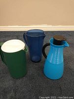 Set of three pitchers/carafe including translucent dark blue plastic pitcher, solid green plastic pitcher with white lid, and bright blue insulated coffee carafe with black handle and lid on carpeted floor.