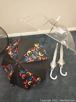 Four umbrellas on display, three clear with white handles uncased, one black with Batman logos.