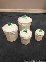 Four white ceramic canisters with octagonal shape and textured raised rose design around the body. Each lid has a green rose-shaped knob.
