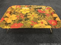 Top view of the rectangular tray showing colorful yellow and orange floral pattern on metal surface, with folded legs holding it elevated.