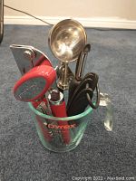 Kitchen utensil set inside Pyrex glass measuring cup showing handles of various utensils including can opener, peeler, ice cream scooper