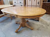 Photo showing the entire oval wooden dining table with pedestal and claw-foot legs, highlighting the carved apron detail and light oak finish.