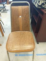 Front-facing vintage vinyl chair with brown upholstery and vertical lighter brown stripe with centered button on backrest, metal tubular frame visible.