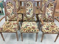 Frontal and angled view of six wooden dining chairs featuring oval wooden detail on backrests and fabric upholstery in floral carpet-style patterned fabric.