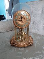 Front view of brass anniversary clock under glass dome showing dial, pillars, pendulum and base