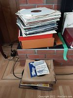 Stack of approximately 50 vintage records in paper sleeves and cardboard boxes on floor by brick fireplace.