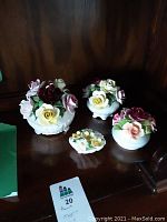 Photo showing four porcelain floral decor figurines on a wooden shelf, different sizes and colors of flowers including reds, yellows and pinks.
