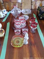 Overview of glasses and dishes on wooden table with chairs in background, showing cranberry rim glasses, clear glass pieces, candy dish on brass stand.