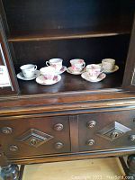 Wide shot of 6 vintage teacups and saucers arranged on a wooden shelf inside a cabinet.