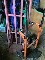 Two dollies side by side, a pink upright hand truck style dolly and an orange convertible dolly with black handles and wheels on a wooden floor.