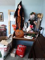 View of 57-inch witch figure in brown-orange dress with black hat, standing next to two male Halloween figurines and a dog figurine on wooden furniture with pumpkin decoration and bags below.