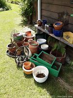 Wide view of assorted outdoor pots and metal baskets arranged on ground and bench outdoors beside a wood wall.