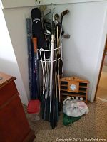 Overall view showing multiple golf clubs in a white metal golf bag carrier, a wooden display shelf with clock, golf balls in bags, and golf umbrellas leaning nearby.