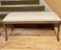 Front view of upholstered padded bench showing beige fabric seat and wooden frame with curved legs.