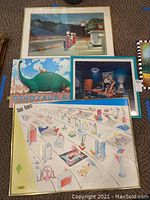 Photo showing four framed whimsical prints laid out on carpet, including 'Gas' by Edward Hopper (depicting a gas station scene), 'Dinosaur Bob' with green dinosaur and rider, 'Dancing Musical Instruments' in a room, and a Bauhaus limited edition print with schematic design.