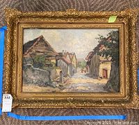 Full view of the painting in gold leaf frame showing village street scene with building facades and foliage.