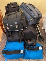 Camera and camcorder bags stacked with tripods and blue sandbags on carpeted floor