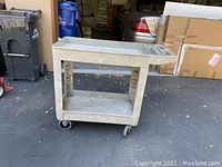 Side angle photo showing entire beige Rubbermaid utility cart with two shelves and caster wheels, with garage background.