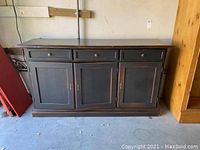 Front view of large black distressed cabinet with three drawers on top and three doors on bottom, showing overall size and condition.