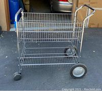 Front view of metal wire cart showing three shelves and handle with two large wheels at the front and two small caster wheels at the back.