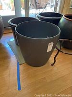 Four black ceramic planters with rounded shape and brown rims, one large gray plastic planter, all grouped on floor